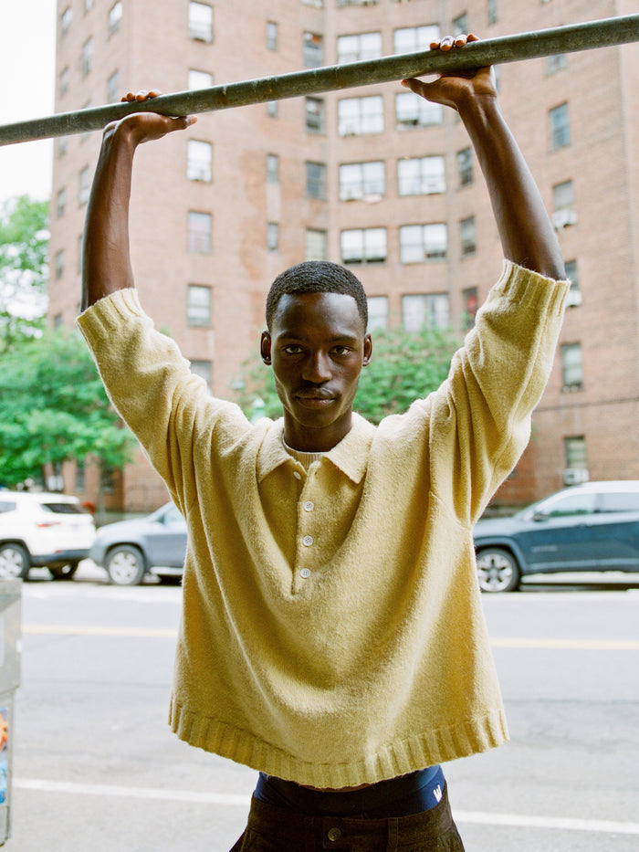 A person wears the FOUND Knit LS Polo Sweater in yellow, shown outdoors holding a metal bar, with brick apartments and parked cars behind them. The sweater is made from soft, brushed knit fabric for comfort and style.