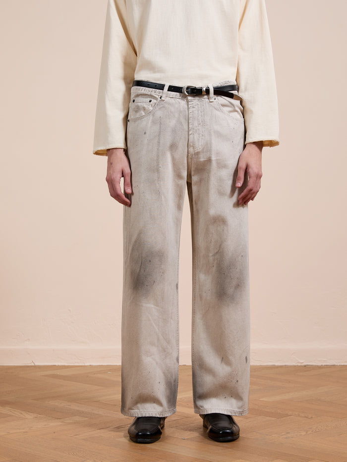 A person wears FOUND's Lacy Baggy Jeans—unisex, relaxed fit, wide-leg, distressed concrete wash with dark stains—paired with a beige long-sleeve shirt, black belt and shoes, standing on a wooden floor against a neutral wall.