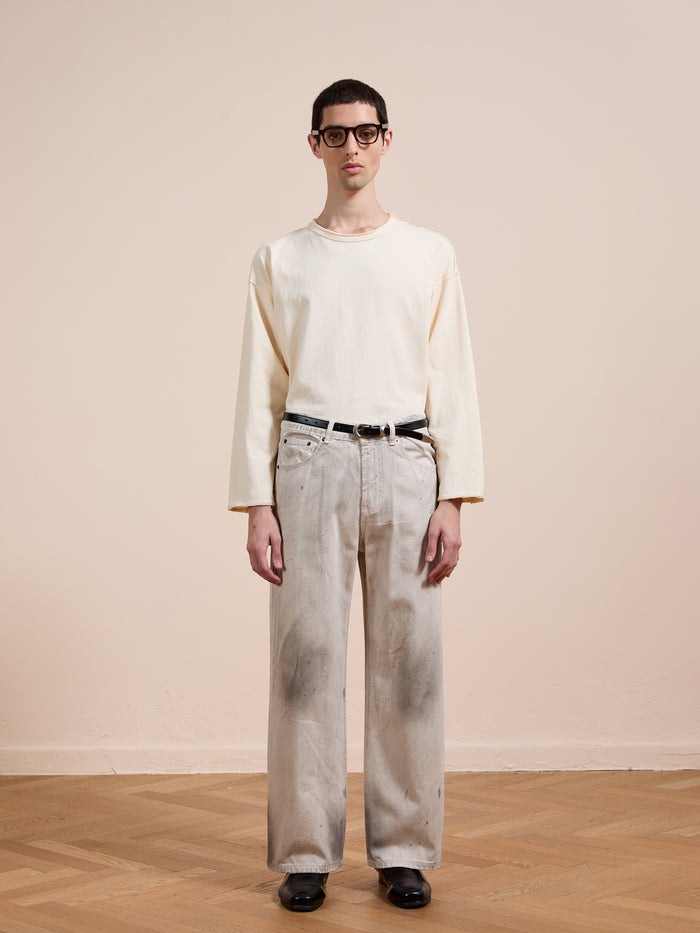 A person stands indoors against a beige wall, wearing glasses, a cream long-sleeve shirt, black belt, black shoes, and FOUND's Lacy Baggy Jeans in a distressed concrete wash.