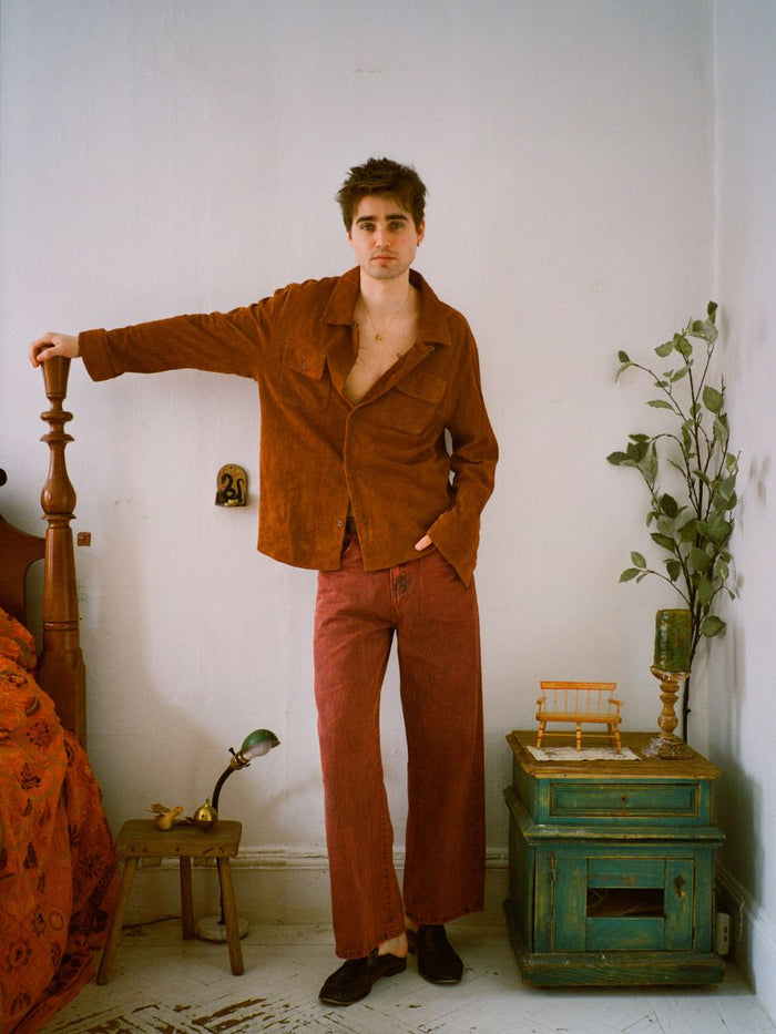 A person in an open brown shirt and FOUND's Lacy Baggy Jeans stands with one hand on a wooden bed frame in a room decorated with a nightstand and various decor.