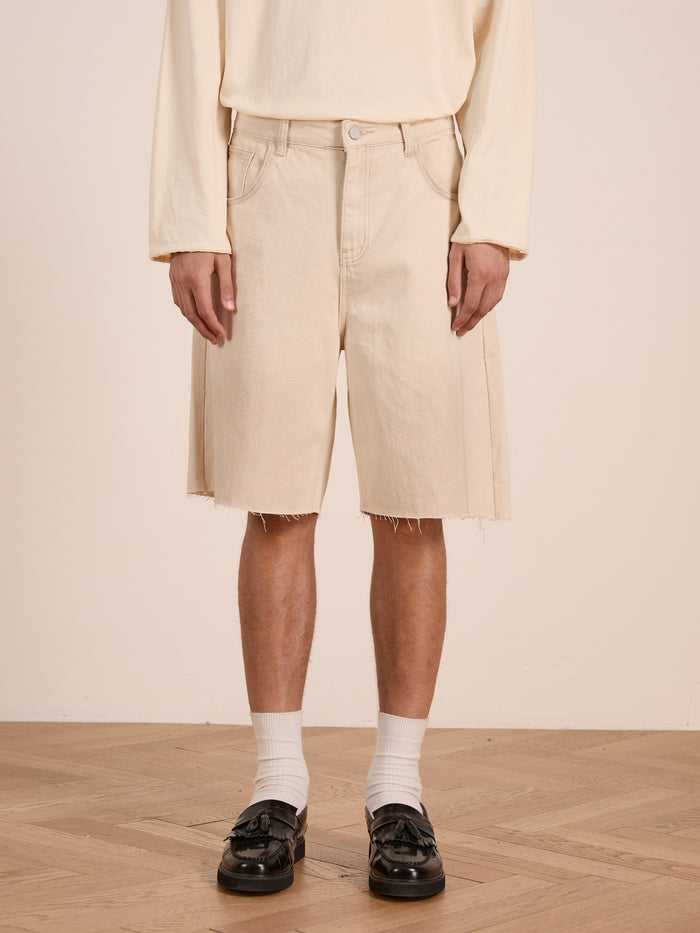 A person wearing FOUND's Lacy Jean Jorts, a long-sleeve beige shirt, white socks, and black loafers stands on a wooden floor against a plain background.