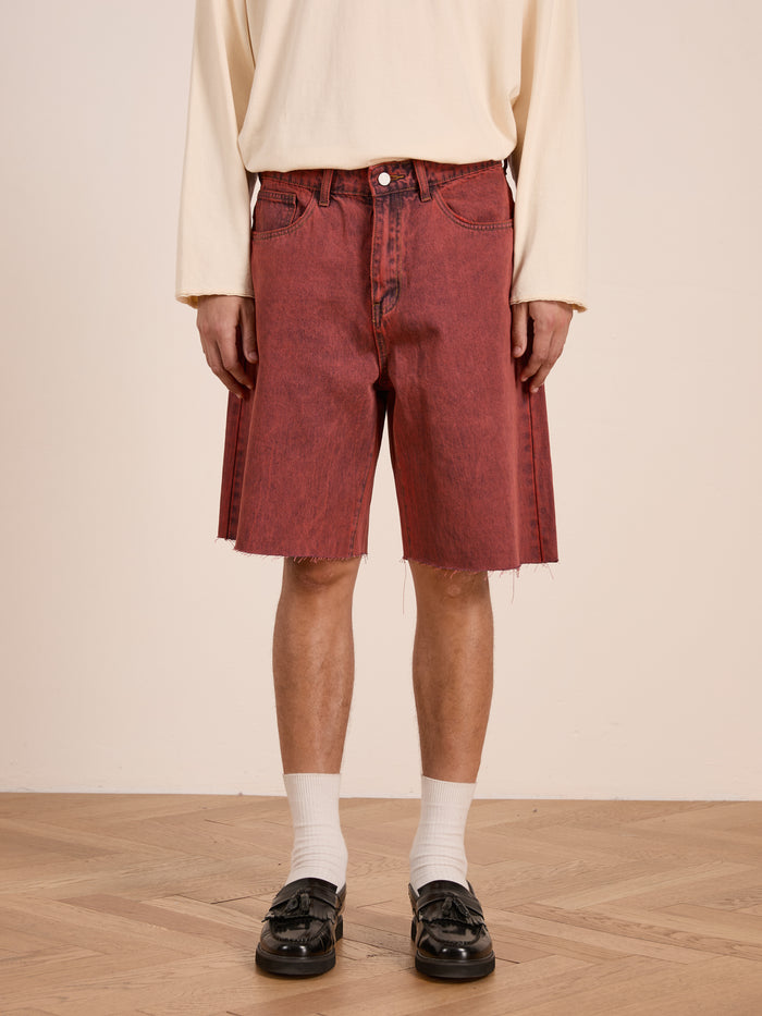 A person wearing a cream long-sleeve shirt, FOUND Lacy Jean Jorts in vintage red wash with frayed hems, white socks, and black loafers stands on a wooden floor.