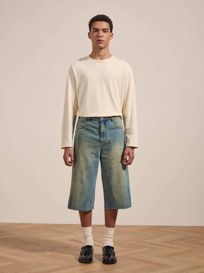 A person stands on a wooden floor wearing a long-sleeve cream shirt, FOUND Lacy Jean Jorts, white socks, and black shoes. The background is plain, and the person looks directly at the camera.