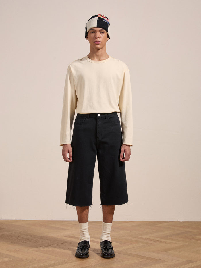 A person stands indoors, wearing a cream long-sleeve shirt, Lacy Jean Jorts by FOUND, black shoes, white socks, and a two-toned beanie. They are looking straight ahead.