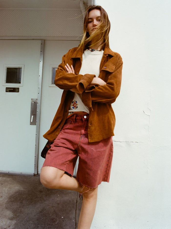 A person with long hair stands against a white wall, wearing a brown jacket, patterned shirt, and FOUND Lacy Jean Jorts in vintage red wash 100% cotton denim, arms crossed and one leg raised.