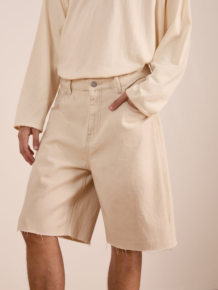 A person models the FOUND Lacy Jean Jorts—distressed denim shorts with a raw hem—paired with a long-sleeve beige shirt, standing with one hand in their pocket.