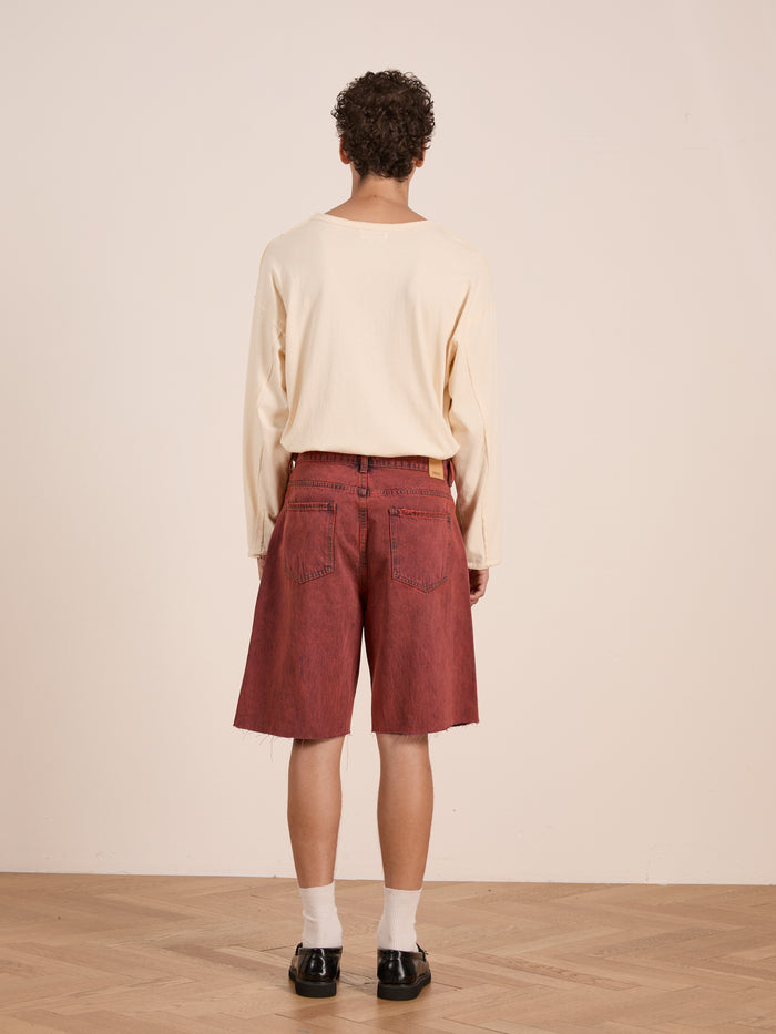 A person with curly hair faces away, wearing a long-sleeve beige shirt, FOUND Lacy Jean Jorts in vintage red wash, white socks, and black shoes on a wooden floor against a plain wall.