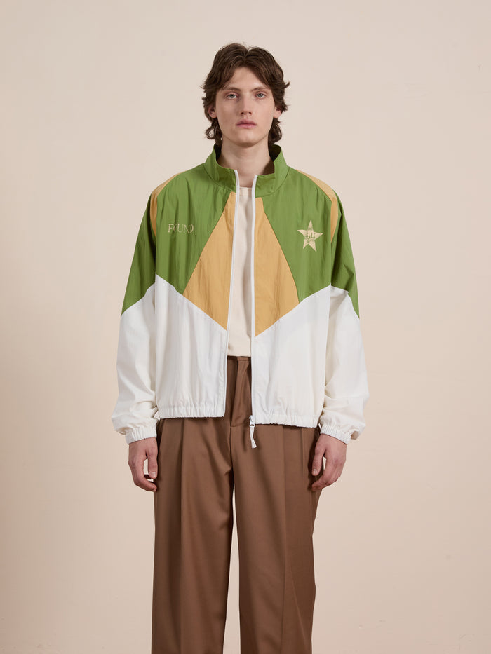 A person in the Legacy Cricket Track Jacket by FOUND, featuring green, white, and tan geometric patterns, stands against a neutral background.