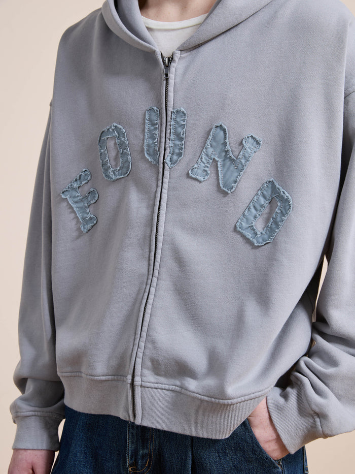A person wearing the FOUND Logo Patch Hoodie, a light gray unisex relaxed-fit zip-up with "FOUND" stitched on the front, styled with dark jeans for a vintage athletic vibe.