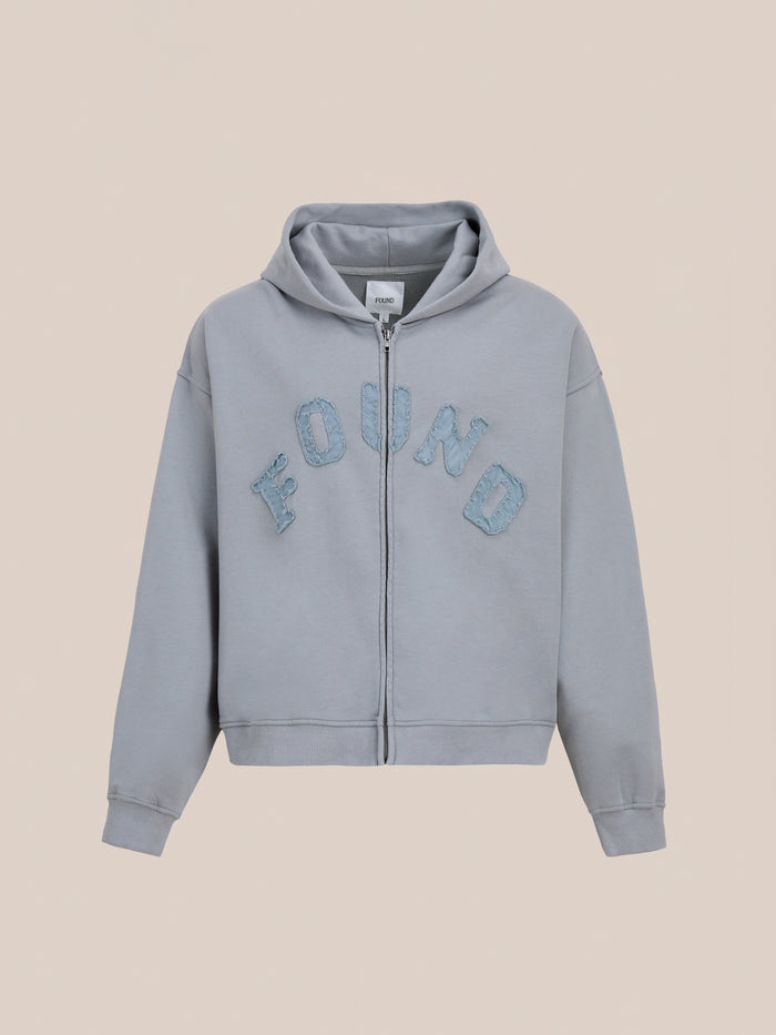 The FOUND Logo Patch Hoodie is a washed gray, unisex zip-up with a hood and bold textured blue "FOUND" lettering on the chest; relaxed fit ideal for vintage athletic style, shown on a plain beige background.