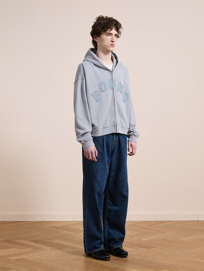 A person stands indoors on a wooden floor in a FOUND Logo Patch Hoodie, unisex relaxed fit blue jeans, and black shoes, against a plain beige wall.