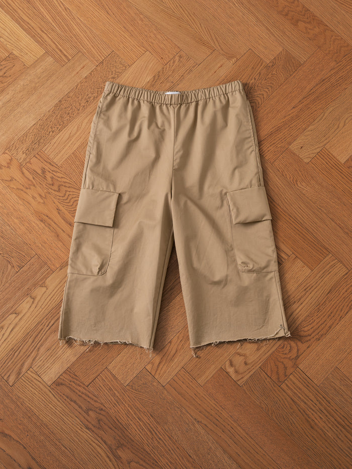 A pair of Long Twill Cargo Shorts by FOUND in beige, featuring frayed hems and made from lightweight cotton fabric, laid flat on a wooden herringbone floor, showcasing the dual cargo pockets.