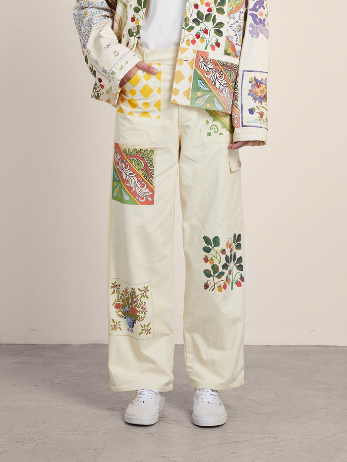 A person in FOUND's Mosaic Pants and an Indo-Aryan patterned jacket with floral and geometric designs stands gracefully on a gray floor, captivating with the vibrant unisex ensemble.