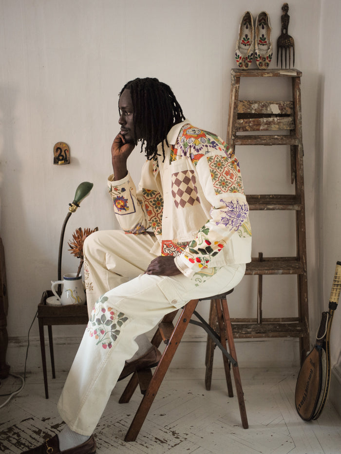 A person with long braided hair sits on a wooden stool, wearing FOUND's Mosaic Pants featuring Indo-Aryan patterns, in a room with a vintage aesthetic, complete with a lamp, ladder, and various decorative items.