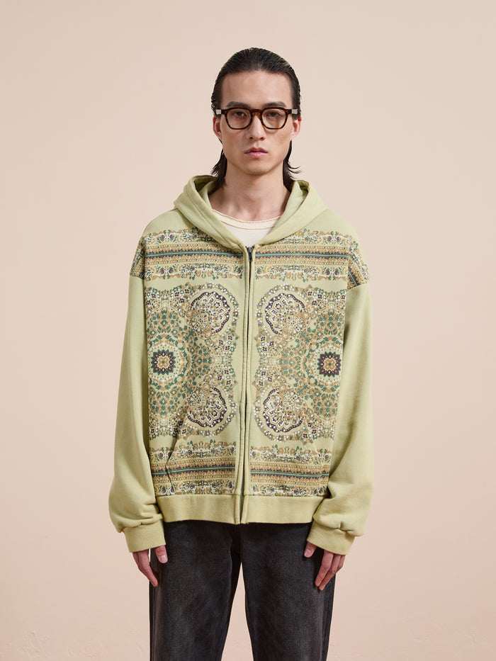 A person with long dark hair and glasses wears the FOUND Motif Zip-Up Hoodie in light green over a white shirt, standing against a beige background—a must-have for streetwear fans.