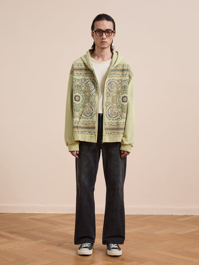Person indoors wearing glasses and a FOUND Motif Zip-Up Hoodie in light green over a white shirt, paired with dark jeans and sneakers, standing against a beige wall and parquet floor.