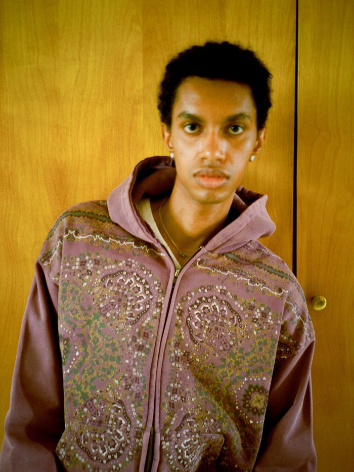 A person with short hair stands against a wooden background, sporting the FOUND Motif Zip-Up Hoodie, a streetwear essential.