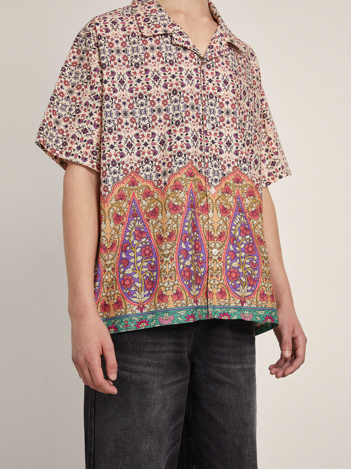A person wearing the FOUND Mughal Camp Shirt, a short-sleeved top featuring vibrant floral and paisley prints, paired with dark jeans.
