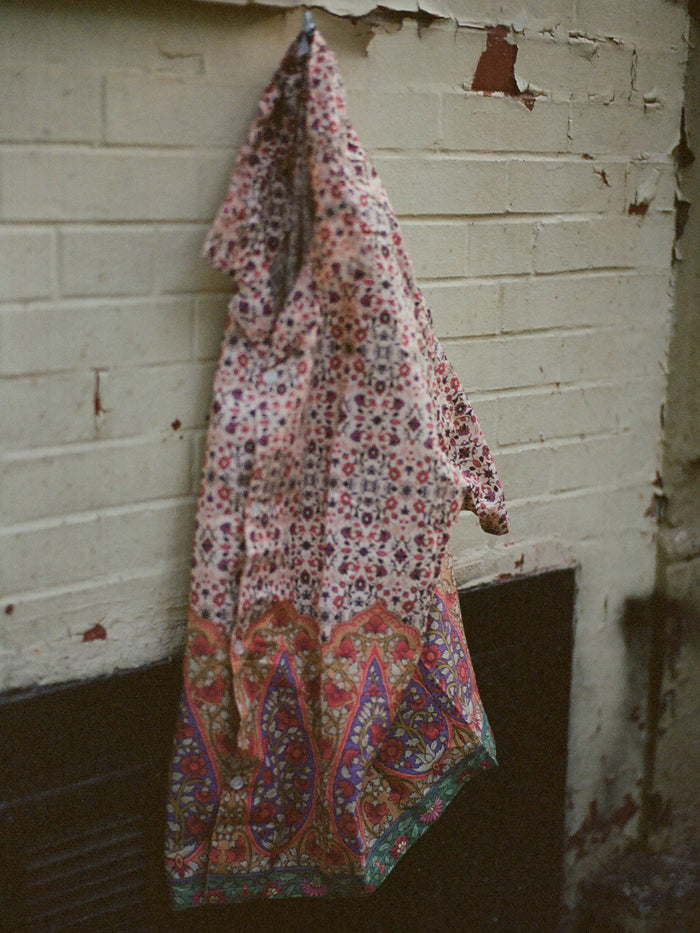 The FOUND Mughal Camp Shirt, adorned with floral and paisley prints, hangs on a hook against a weathered pale brick wall.