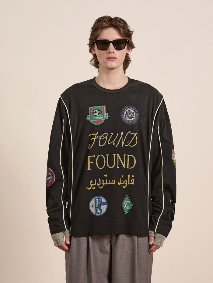 Wearing the unisex Multi Language LS Jersey by FOUND, a person is styled in casual sunglasses and a black long-sleeve shirt featuring various patches and "FOUND" repeated, creating a retro look against a plain backdrop.