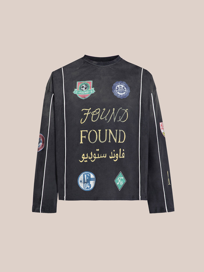 The Multi Language LS Jersey by FOUND is a black long-sleeve jersey adorned with patches and gold "Found" with Arabic text on the front, reflecting a retro football kit style.
