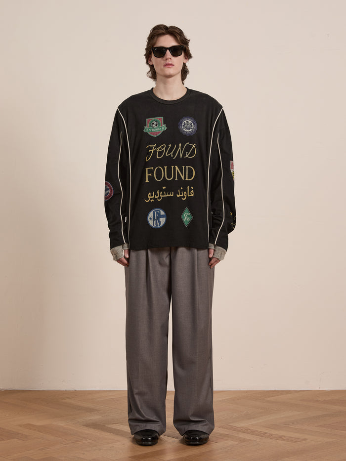 A person embodies unisex casual wear in a retro style, wearing sunglasses and the "FOUND" Multi Language LS Jersey in black, adorned with various patches. They pair it with gray wide-leg pants and black shoes while standing on a wooden floor against a plain wall.