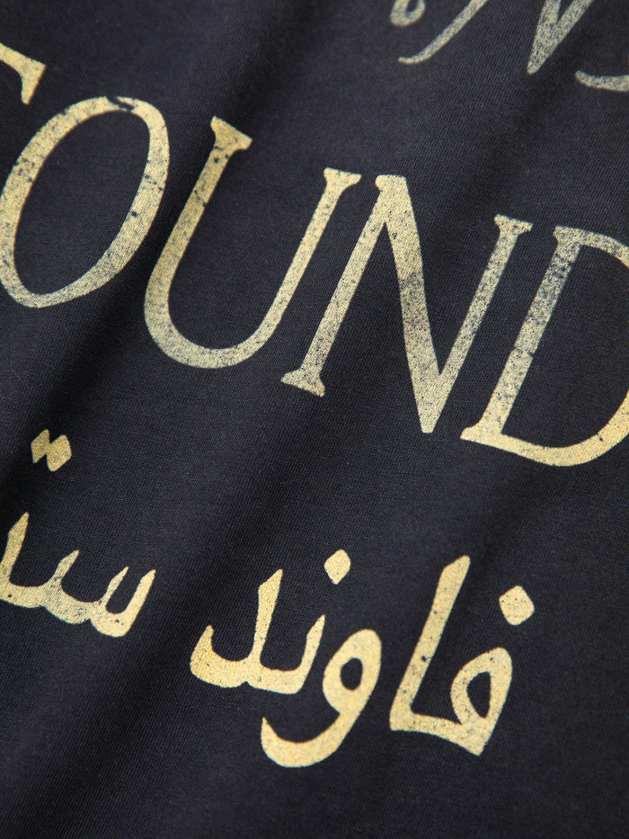 Close-up of a black fabric with a retro design, showcasing "FOUND" in worn gold lettering and Arabic script below—ideal for lovers of unisex casual wear with a vintage touch. This is the Multi Language LS Jersey by FOUND.