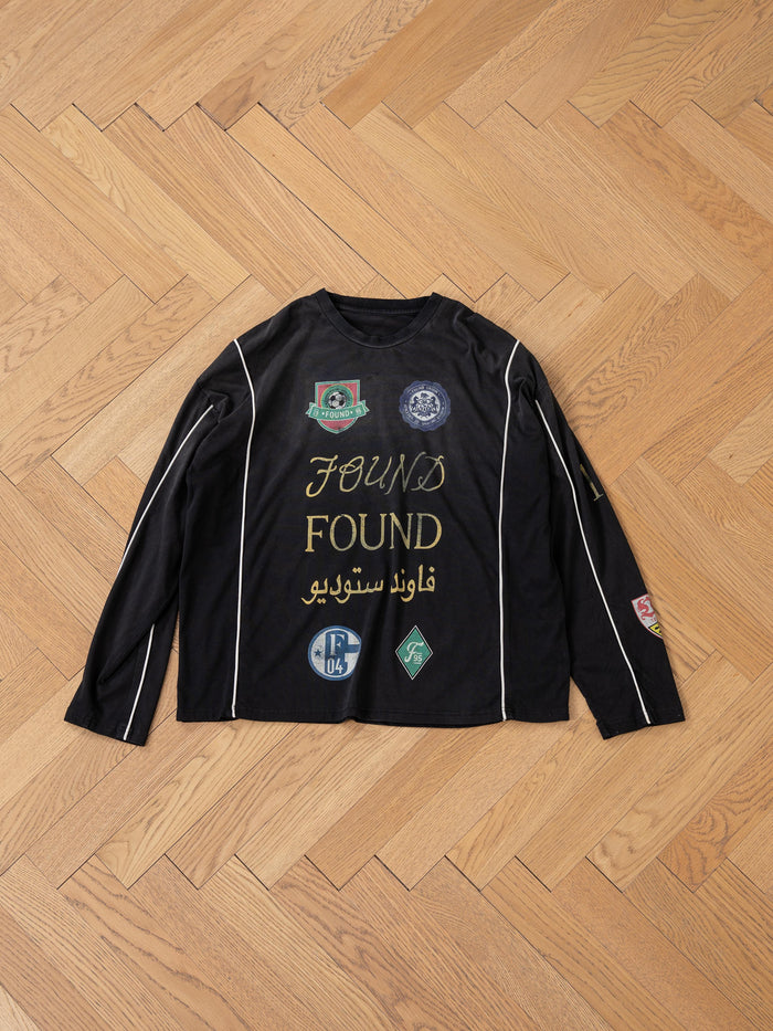 The FOUND brand's "Multi Language LS Jersey" is a retro unisex black long-sleeve shirt adorned with white text and various patches on both sides, elegantly displayed on a herringbone wood floor, combining classic style with modern flair.