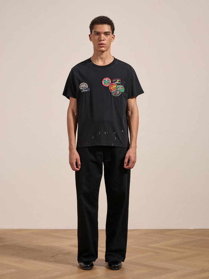 A person stands against a plain background, wearing the FOUND Multi Patch Washed Tee with various colorful patches on the front and black pants.