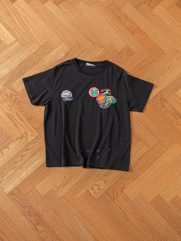 The Multi Patch Washed Tee by FOUND, featuring a black design adorned with multiple colorful patches, is displayed on a wooden parquet floor. The shirt has small holes near the bottom.