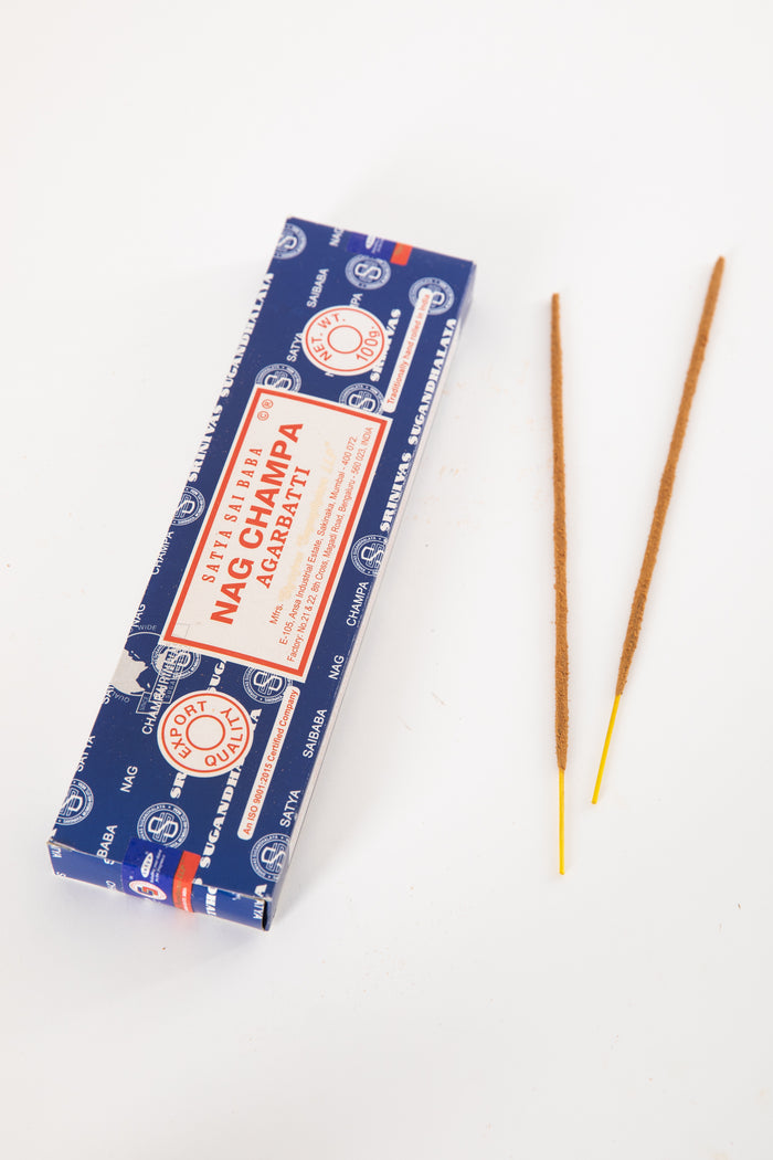 A box of Satya Sai Baba Nag Champa incense with two incense sticks placed in front of it on a white background.