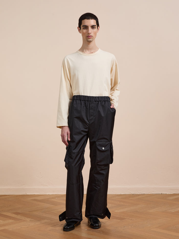 Indoors against a plain beige background, a person wears FOUND Nylon Cargo Pants in black with pockets, paired with a long-sleeve cream shirt and black shoes.