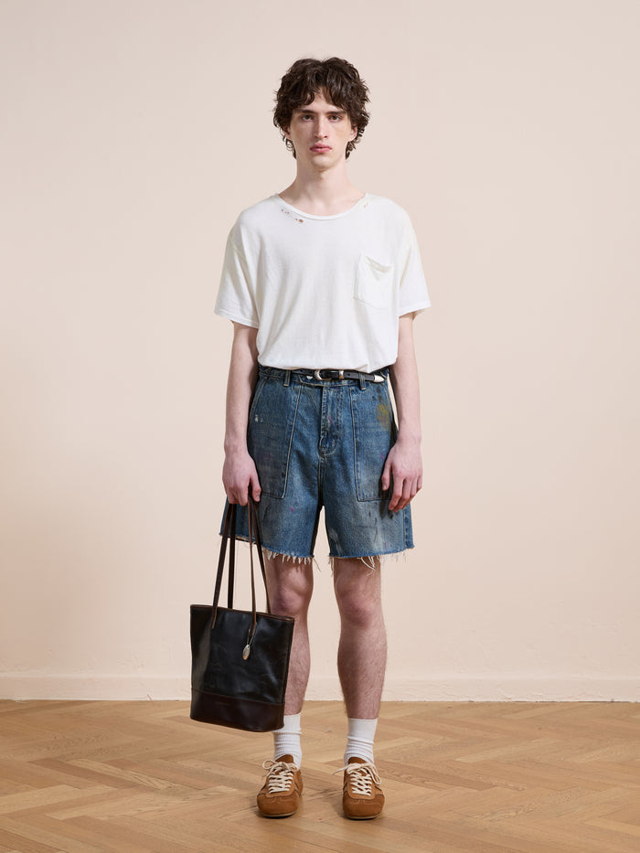 A person stands indoors against a plain background wearing a white t-shirt, Found Paint Studio Denim Shorts, white socks, brown shoes, and holding a black tote bag.