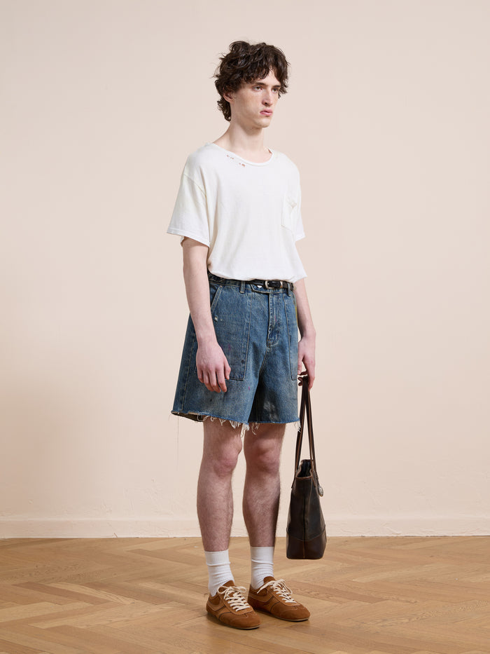 Indoors against a plain beige background, a person wears a white t-shirt, Found Paint Studio Denim Shorts, white socks, brown shoes, and holds a dark handbag.