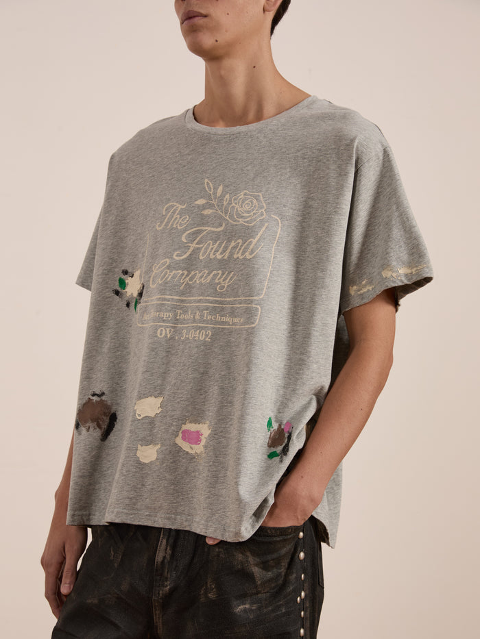 A person stands against a plain backdrop wearing a gray oversized "Painters Company Tee" by FOUND, featuring vintage-style paint splatters. Hands in pockets, the unisex look effortlessly embodies workwear-inspired style.