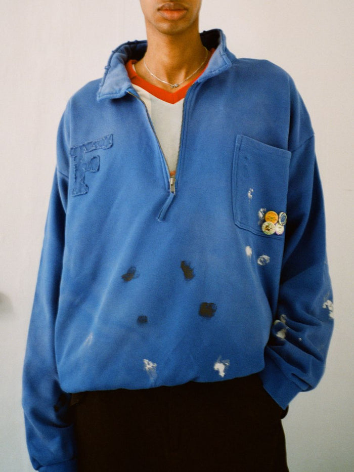 Against a plain backdrop, someone wears the FOUND Painters Pullover Sweatshirt. It's an oversized blue half-zip made of 100% cotton, with worn fabric and button pins enhancing its casual painter's charm.
