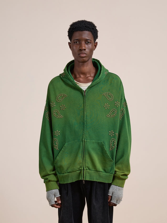 A person wearing the FOUND Paisley Studded Zip Up Hoodie in green, featuring a unisex relaxed fit, pairs it with a white shirt underneath, fingerless gray gloves, and black pants while standing against a plain beige background.