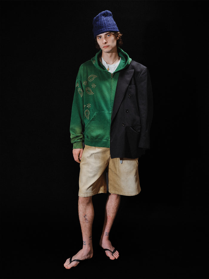 A person wearing a blue beanie, the FOUND Paisley Studded Zip Up Hoodie in green, a black blazer, beige shorts, and flip-flops stands against a black background.