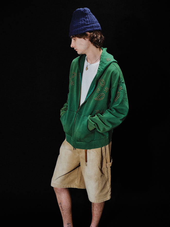 A person wearing the FOUND Paisley Studded Zip Up Hoodie in vintage green, along with a white t-shirt, beige shorts, and a blue knit hat, stands with hands in pockets against a black background.