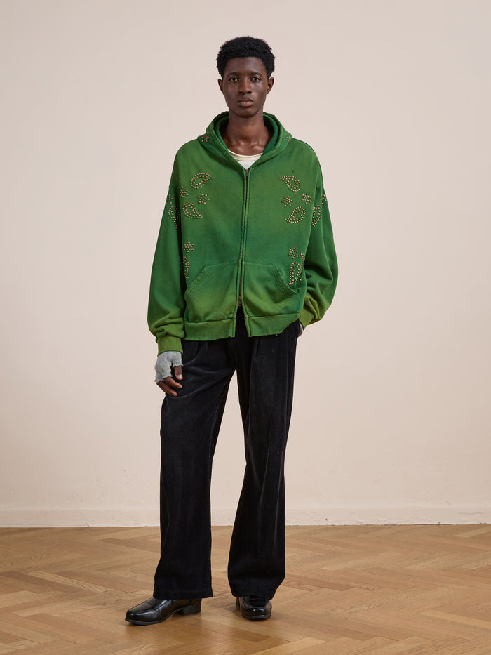 A person indoors sports the FOUND Paisley Studded Zip Up Hoodie—green with decorative patterns—paired with black wide-leg pants, black shoes, and a gray fingerless glove for a cool, vintage look.