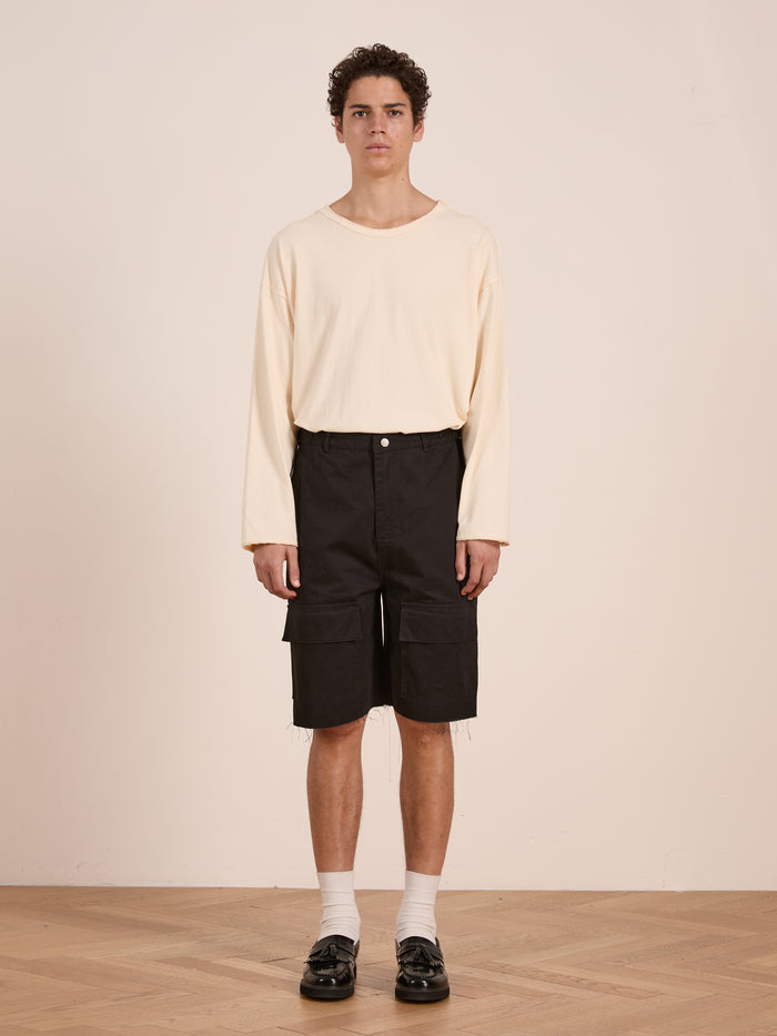 Indoors, a person wears a long-sleeve cream shirt, Found Parachute Cargo Twill Shorts in black, white socks, and black shoes.