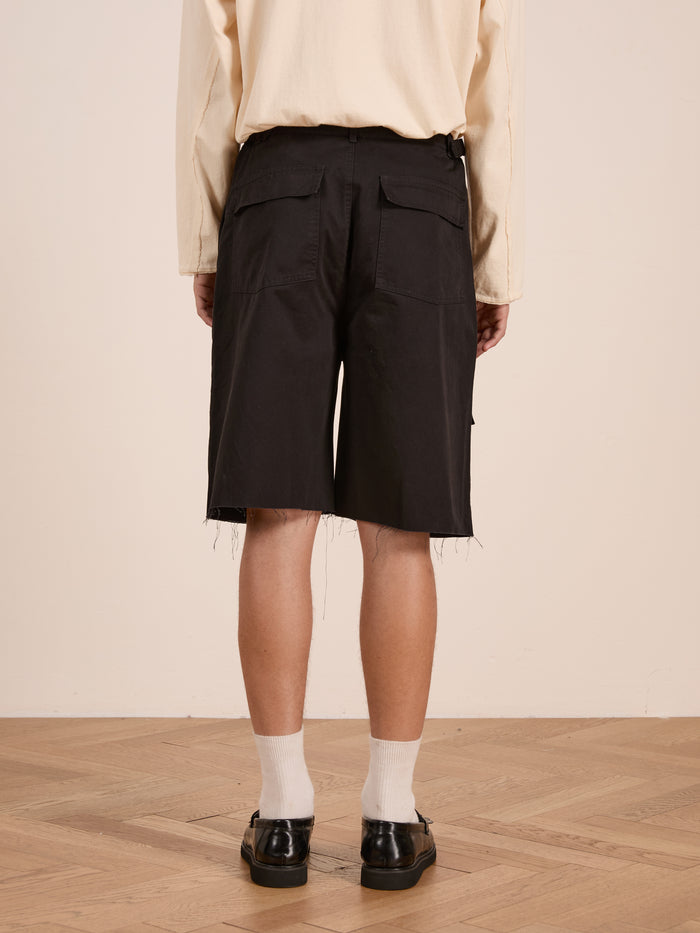 Person standing on a wooden floor, shown from the back, wearing a cream long-sleeve shirt, white socks, black shoes, and Found Parachute Cargo Twill Shorts—black with frayed hems for a utility-inspired look.