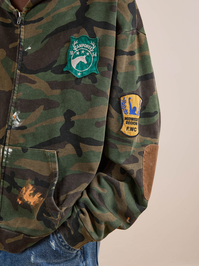 Wearing the FOUND Patch Camo Zip Up Hoodie, a relaxed fit unisex style featuring a green "Camporee 1954" patch on the chest and a yellow "Midwest Region F.W.C." patch on the sleeve.