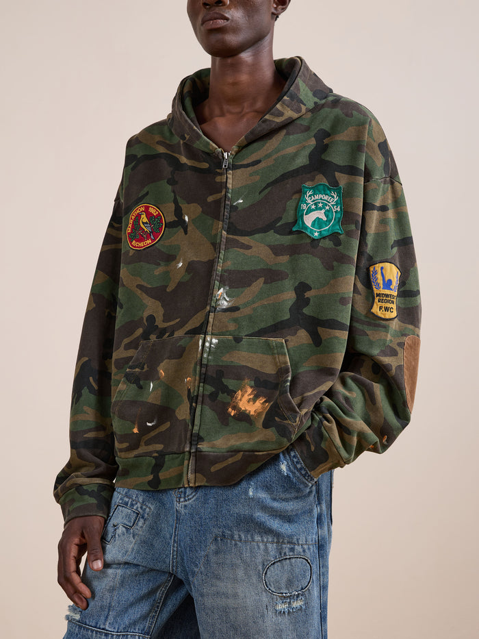 A person wears the FOUND Patch Camo Zip Up Hoodie, styled with relaxed fit blue jeans that have visible stitching and a knee patch.