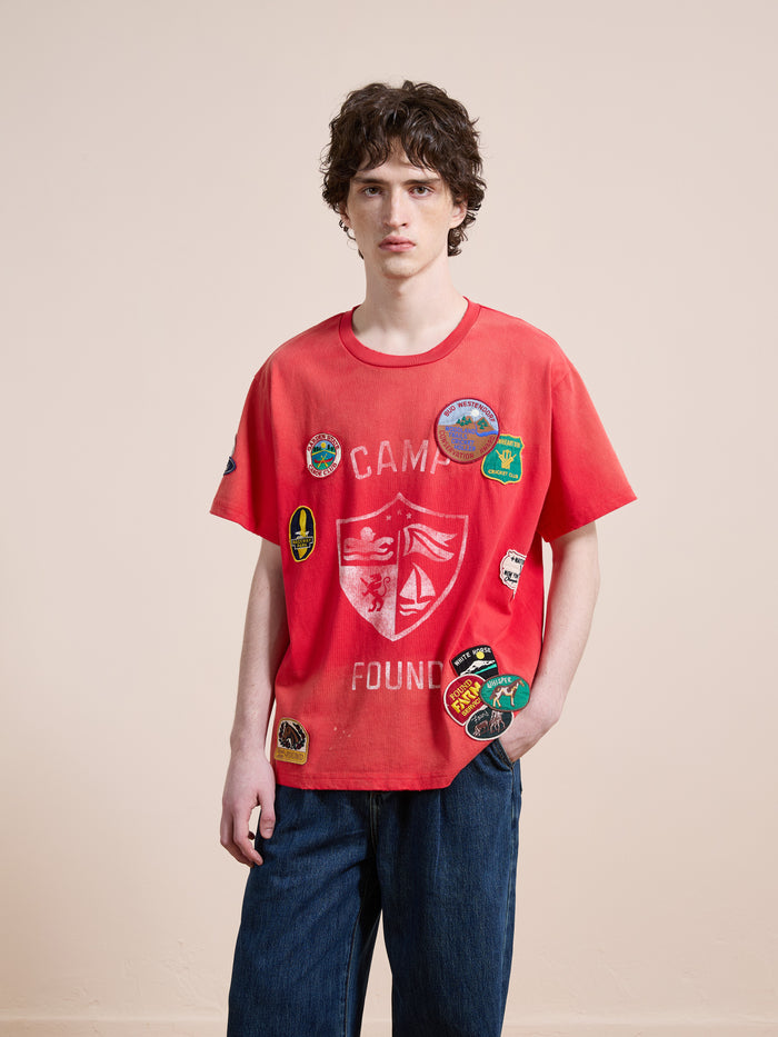 A young man with curly hair wears the FOUND Patchwork Camp Tee and blue jeans, standing against a plain beige background.
