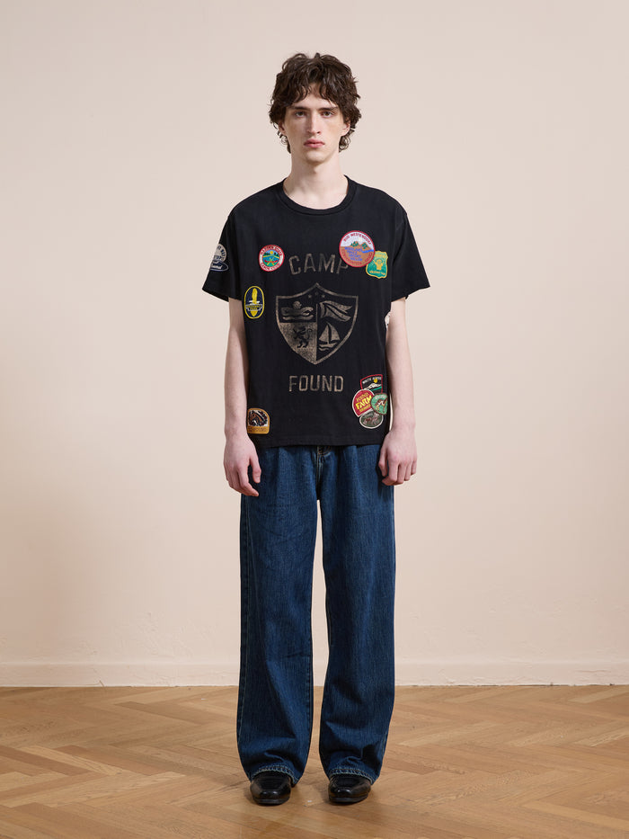 A person stands indoors against a plain wall, wearing the FOUND Patchwork Camp Tee and wide-leg blue jeans for a cool unisex relaxed look.