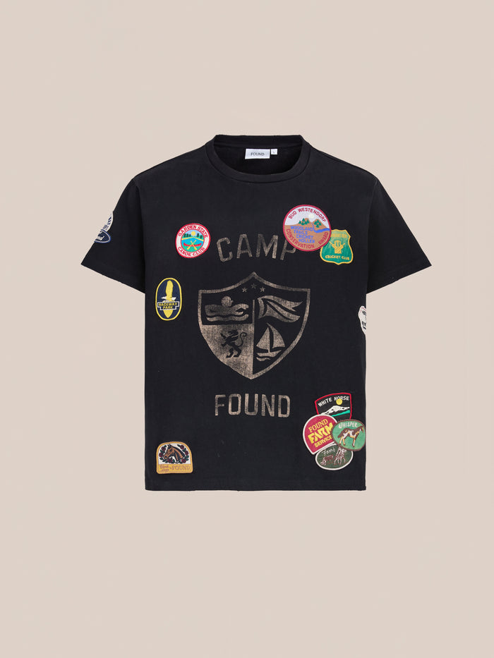 The Patchwork Camp Tee by FOUND is a vintage black unisex relaxed-fit shirt, showcasing a crest and “Camp Found” text with colorful embroidered patches on the front and sleeves.