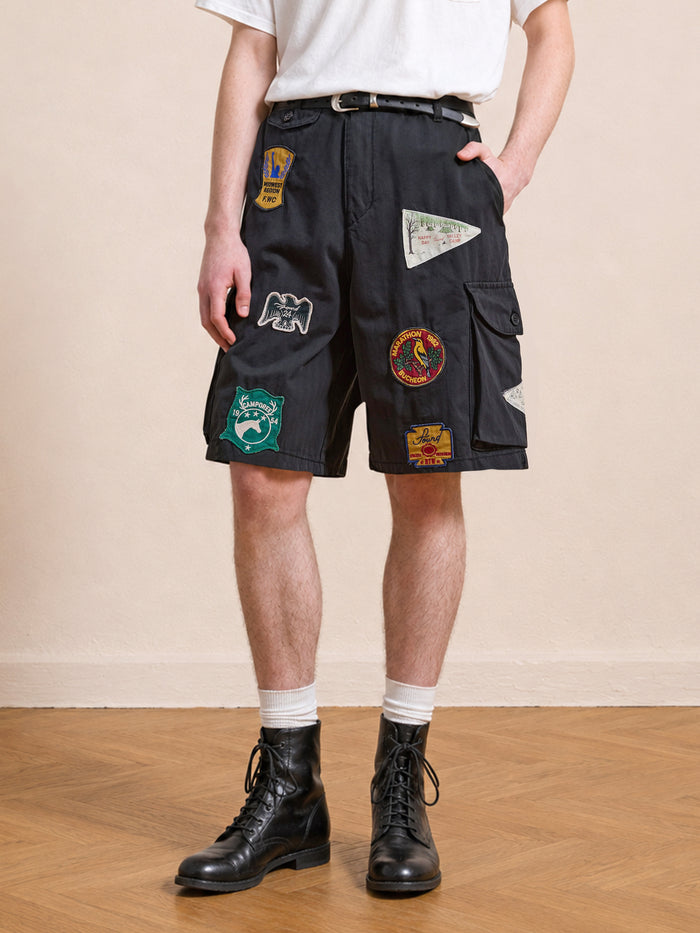 Someone wearing Found Patchwork Cargo Shorts in vintage black with colorful patches, paired with a white t-shirt, white socks, and black lace-up boots, standing on a wooden floor.