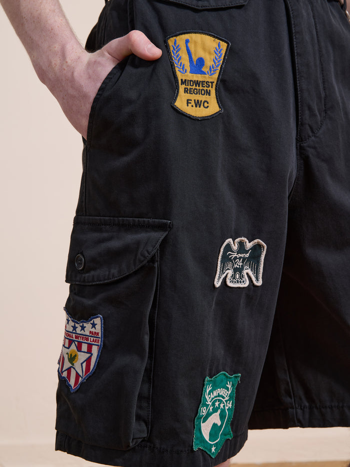 A person wears Found Patchwork Cargo Shorts, a unisex relaxed fit style in vintage black wash, detailed with embroidered logo patches on the front and side pocket.