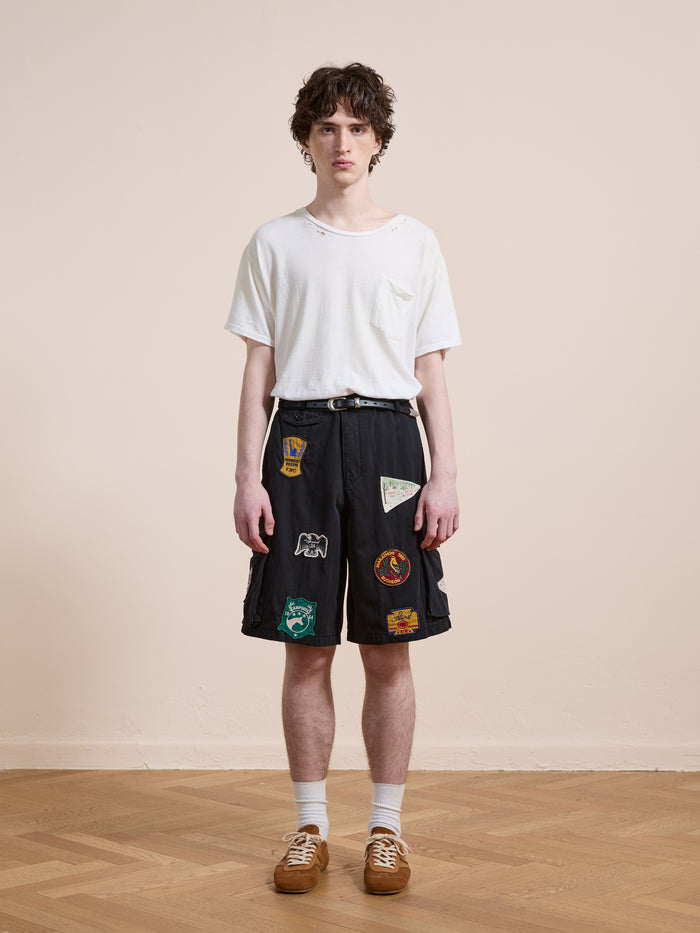 A young person stands indoors facing forward against a plain background, wearing Found's Patchwork Cargo Shorts in a vintage black wash with a relaxed unisex fit, paired with a white t-shirt, white socks, and brown shoes.